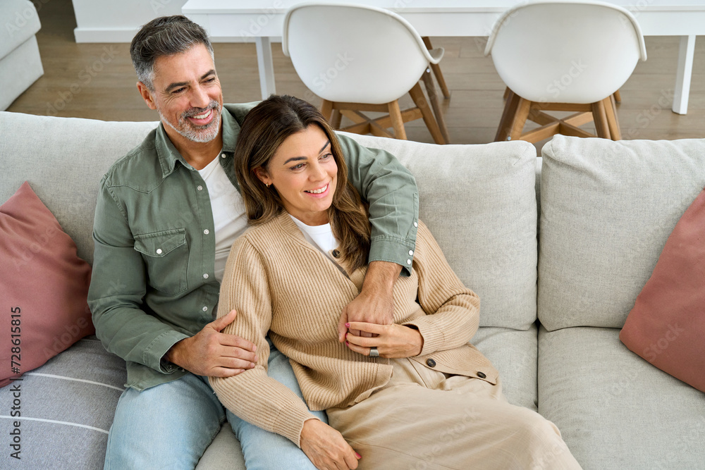 Happy middle aged mature couple hugging looking away relaxing on couch at home. Relaxed serene older man and woman in love sitting on sofa together embracing in modern house living room. Happy middle aged mature couple hugging looking away relaxing on couch at home. Relaxed serene older man and woman in love sitting on sofa together embracing in modern house living room.