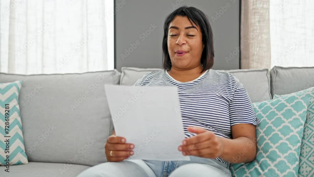 A middle-aged hispanic woman at home reacts with various emotions while reading a document on a couch, enhancing this typical indoor scene.
