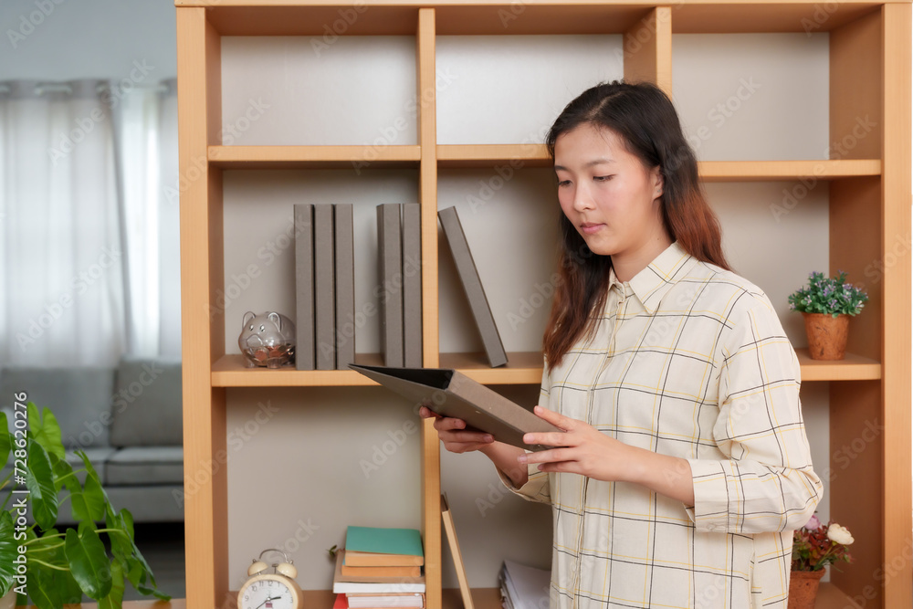 Concentrated Asian woman reads from a folder, standing in a home office with modern wooden shelves. Business-casual attire and a focused expression, Asian female stands reading in neat workspace Concentrated Asian woman reads from a folder, standing in a home office with modern wooden shelves. Business-casual attire and a focused expression, Asian female stands reading in neat workspace