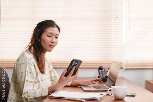 Engaged in work, Asian businesswoman interacts with smartphone, balancing tech in corporate role. Multitasking with ease, professional uses her phone alongside laptop in dynamic work environment