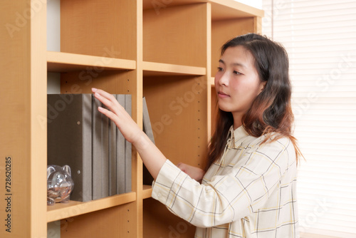 Asian office worker selects a folder on a shelf, surrounded by a warm wooden interior and soft lighting. thoughtful Asian professional reaches for a file in a well-organized, sunlit office space