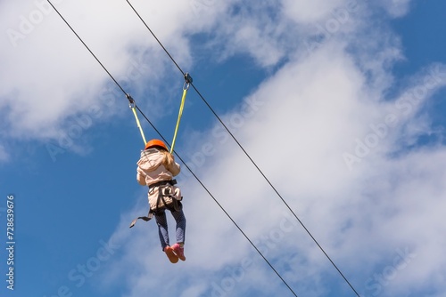 Child girl practicing zip lining outdoors. In the background blue sky with white clouds.
