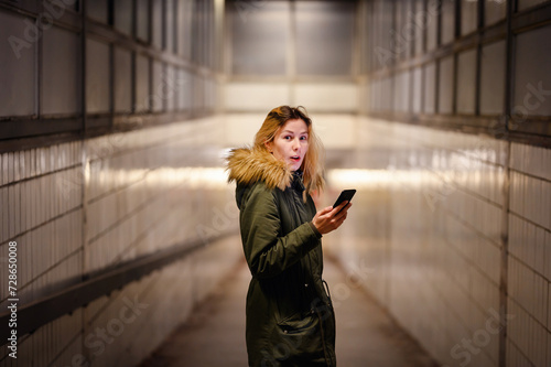 A woman shows a surprised expression while looking at her phone in a subway passage