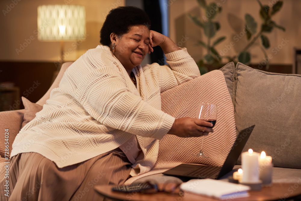 Fototapeta premium Side view portrait of senior Black woman clinking wine glass to screen during video chat with friend or long distance partner copy space