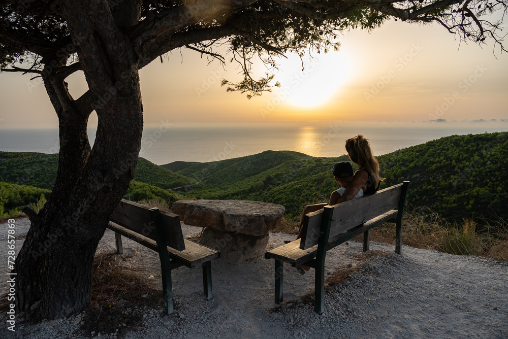 Family sitting on bench and looking at sunset.  Sunset pine trees, olive groves and sea view. Mediterranean sunset from the Agalas viewpoint. Sitting and watching sunset in Zakynthos, Greece.