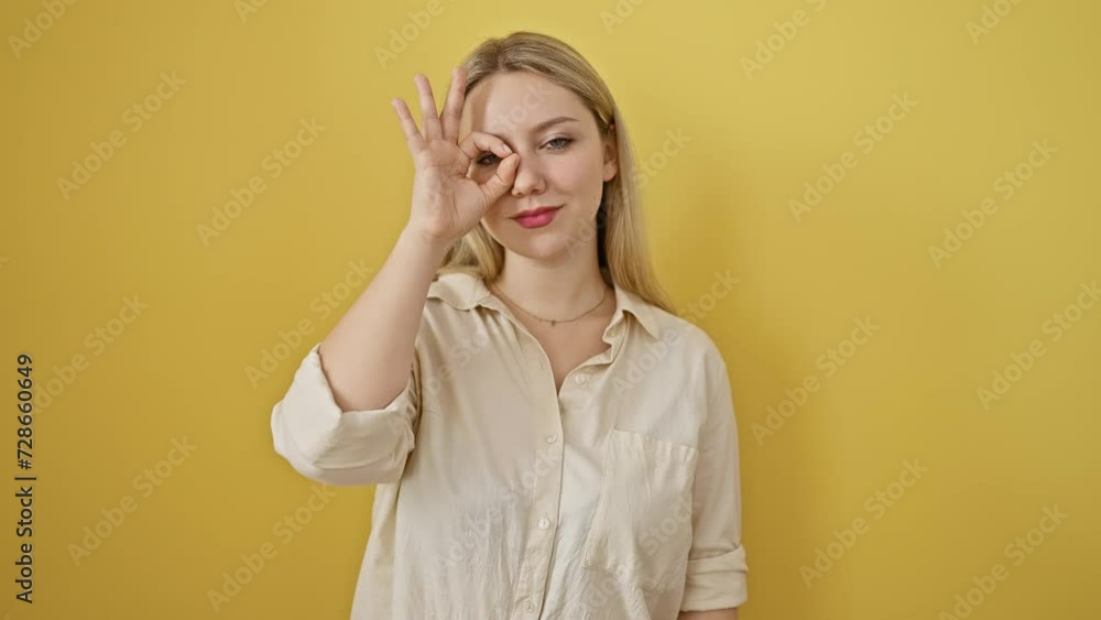 Joyful young blonde woman confidently grinning, standing isolated on a yellow background. with a happy face expression, she uses her fingers to create a-okay symbol by looking through her finger. Joyful young blonde woman confidently grinning, standing isolated on a yellow background. with a happy face expression, she uses her fingers to create a-okay symbol by looking through her finger.