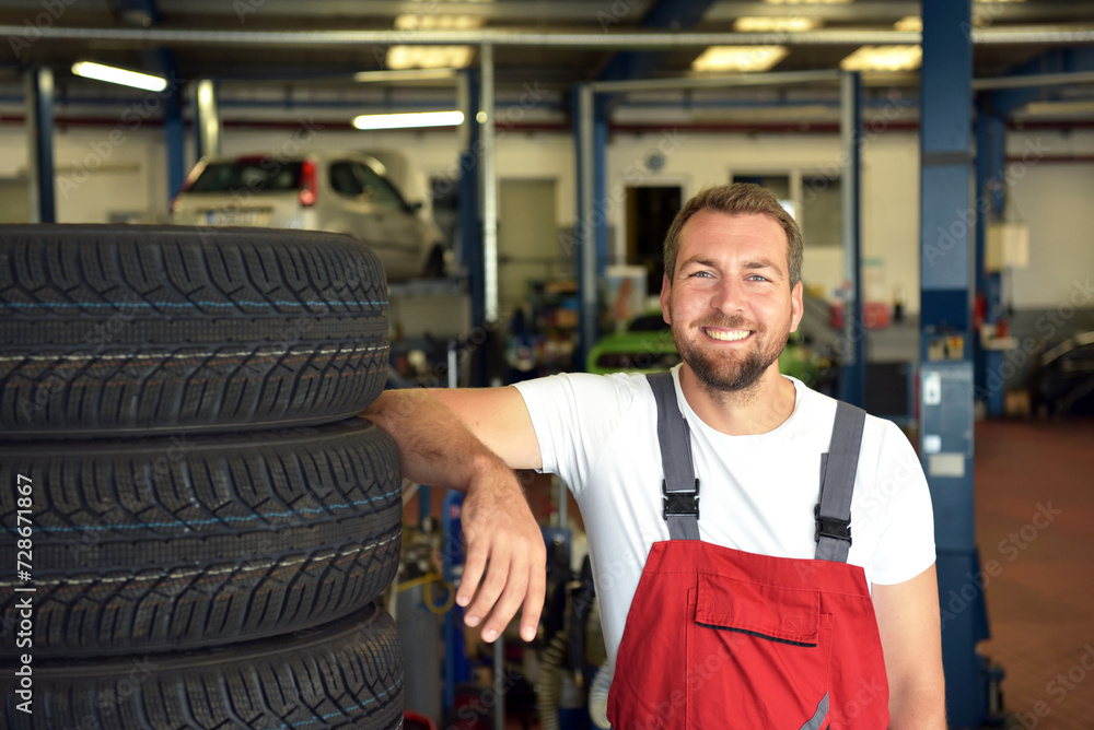 Portrait of successful smiling car mechanic in a workshop on a stack of tires at his workplace - obrazy, fototapety, plakaty 