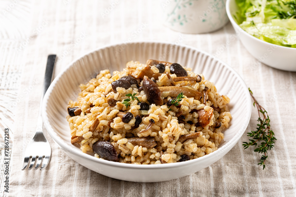 Mushroom barley risotto or orzotto in a white plate on a line tablecloth. Made with pioppini and porcini mushrooms. Green salad on background. Italian Vegan food. Mushroom barley risotto or orzotto in a white plate on a line tablecloth. Made with pioppini and porcini mushrooms. Green salad on background. Italian Vegan food.