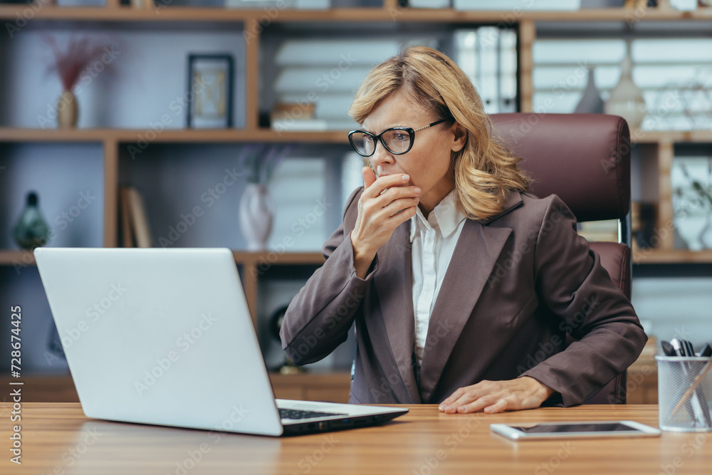 Shocked senior business woman sitting in the office at the desk and looking worriedly at the laptop screen. Problems with accounts, received bad news in the mail, reads the message. Shocked senior business woman sitting in the office at the desk and looking worriedly at the laptop screen. Problems with accounts, received bad news in the mail, reads the message.