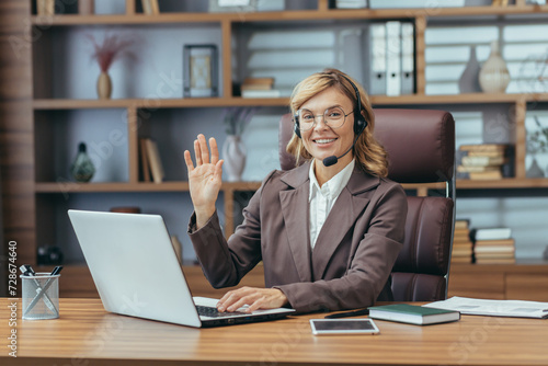 Portrait of a smiling and successful senior business woman working in the office with a laptop, wearing a headset, looking smugly at the camera, waving and greeting. photo