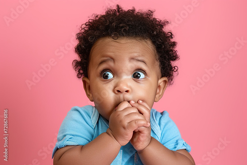 Scared African American baby boy and biting nails in studio with oops reaction on pink background