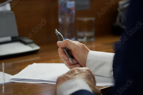 An elderly man in a business suit with cuffs, sitting at a desk with pen and documents. Concept of writing a will, drafting a contract or filling out forms. Photo. No face. Selective focus photo