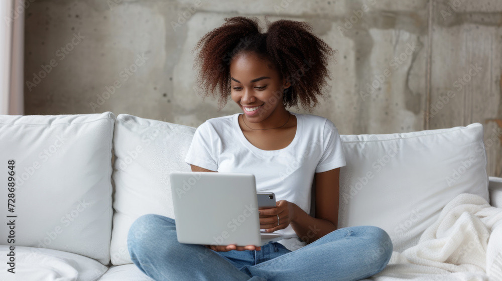 Naklejka premium smiling young woman is comfortably seated on a couch, looking at a tablet while holding a smartphone in her other hand