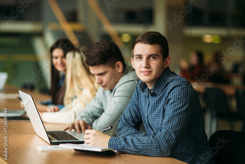 Group of college students studying in the school library, a girl and a boy are using a laptop and connecting to internet. Boy look to the camera