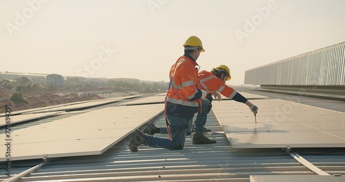 Two engineer workers in safety gear install solar panels on a rooftop, with a suburban landscape stretching out in the distance photo