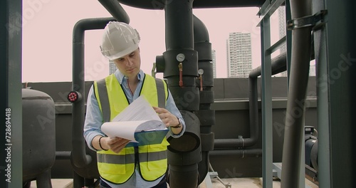 Engineer in hard hat and reflective vest inspecting paperwork at an urban industrial site examining a document on a rooftop with industrial pipes and valves in the background