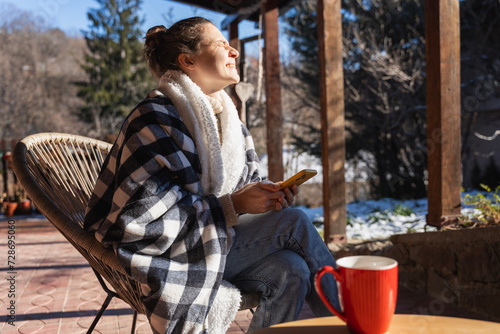 Young woman in a warm blanket on the terrace of a country house cottage enjoying the early spring sun, with a cup of coffee and a smartphone