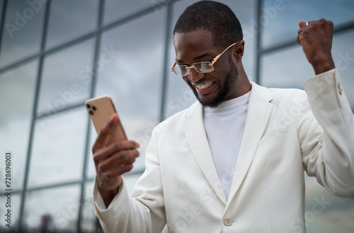 Excited black African American male holding and looking at his phone whit a bussiness building in the background