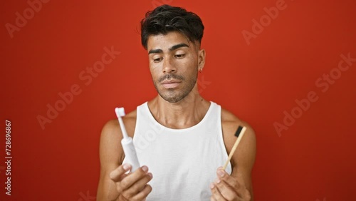 Pensive young man comparing traditional toothbrush with electric one against red backdrop. photo