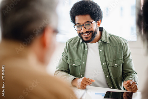 African-American man with a warm smile, wearing glasses and a green casual shirt, is engaged in conversation with an out-of-focus colleague, suggesting an approachable and friendly office discussion photo