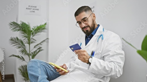 A bearded man in glasses and a white coat checks his smartphone and credit card in a bright medical office. photo