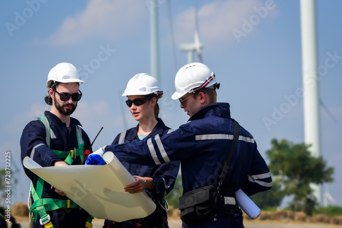 male and female electrical engineers and technicians wearing safety uniform workwear working outdoor, look at blueprints and discuss on a wind farm or wind turbines field used to produce electricity.
