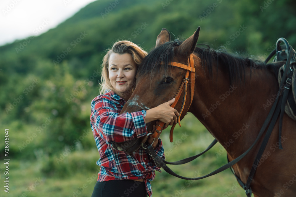 Obraz premium Happy blonde with horse in forest. Woman and a horse walking through the field during the day. Dressed in a plaid shirt and black leggings.