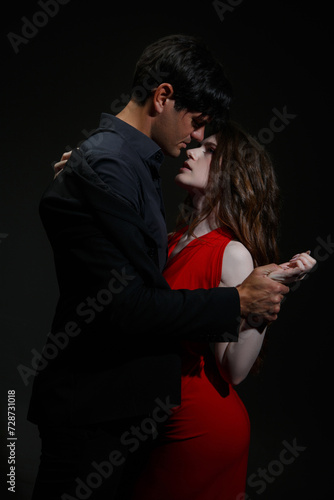 A passionate kiss between a man in a black suit and a woman in a red dress on a dark background.