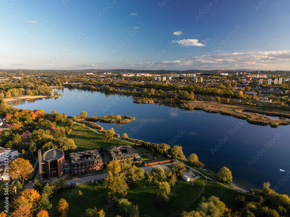 Fototapeta premium Aerial view of Bagry lake and autumn forest in Poland. Amazing panoramic landscape with waters of spectacular Bagry lagoon and road along beach, houses of town and scenic tops of trees under blue sky
