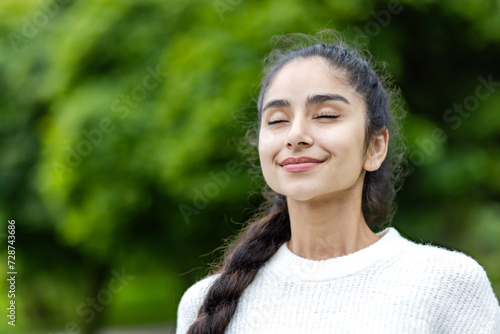 A young beautiful Indian woman is standing outside in a park in a white sweater and with her eyes closed is taking deep breaths and enjoying herself. Close-up photo