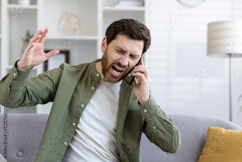 Vászonkép Mad unshaved man talking by cellphone and shouting angrily while raising hand in living room