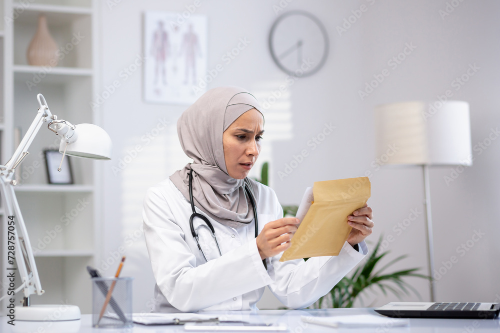 Disturbed female nurse in hijab opening paper envelope while sitting at workplace. Disappointed hospital worker reading unpleasant message from laboratory with unsuccessful treatment results.