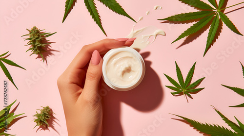 Hand holds natural cannabis cream in jar on pink background with hemp leaves. Natural ingredients cosmetics concept. Organic CBD skincare lotion with fresh marijuana buds on light pink