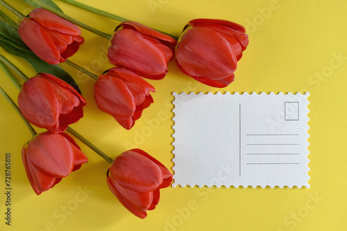 Bouquet of fresh red tulips and greeting card on yellow background. Top view, mock up. Close-up. Copy space. Selective Focus.