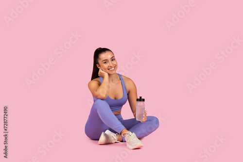 Young woman in sportswear with water bottle, smiling on pink background