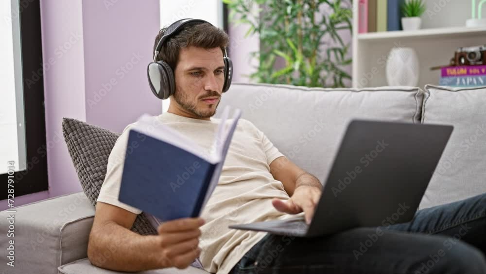 A focused man with headphones reads while multitasking on a laptop in a cozy living room. A focused man with headphones reads while multitasking on a laptop in a cozy living room.