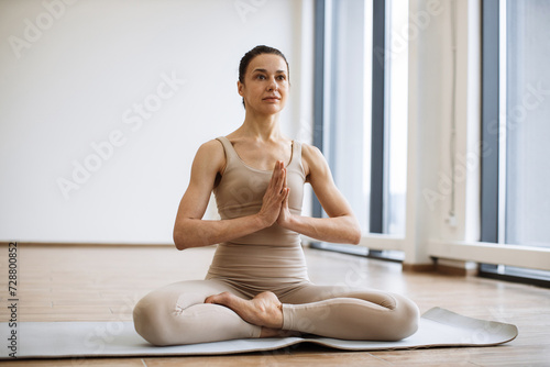 Front view of caucasian lady in beige athletic wear doing Padmasana exercise on rubber mat in bright fitness studio. Yoga practitioner improving concentration with namaste gesture in lotus pose.