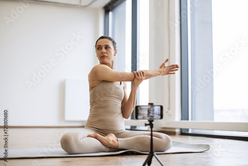 Sporty adult doing padmasana exercise on mat shooting video content on smartphone during yoga class. Side view of senior lady blogger in sportswear meditating in cross-legged sitting pose in studio.