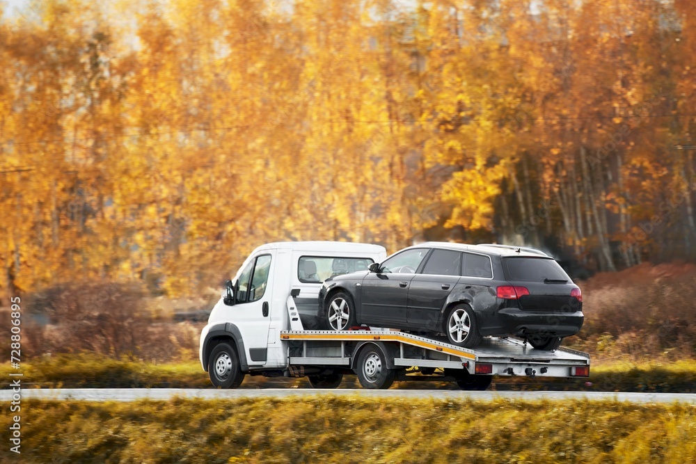 A rollback tow truck transports a broken car on the public road. Car failure during the journey. Roadside assistance in action.