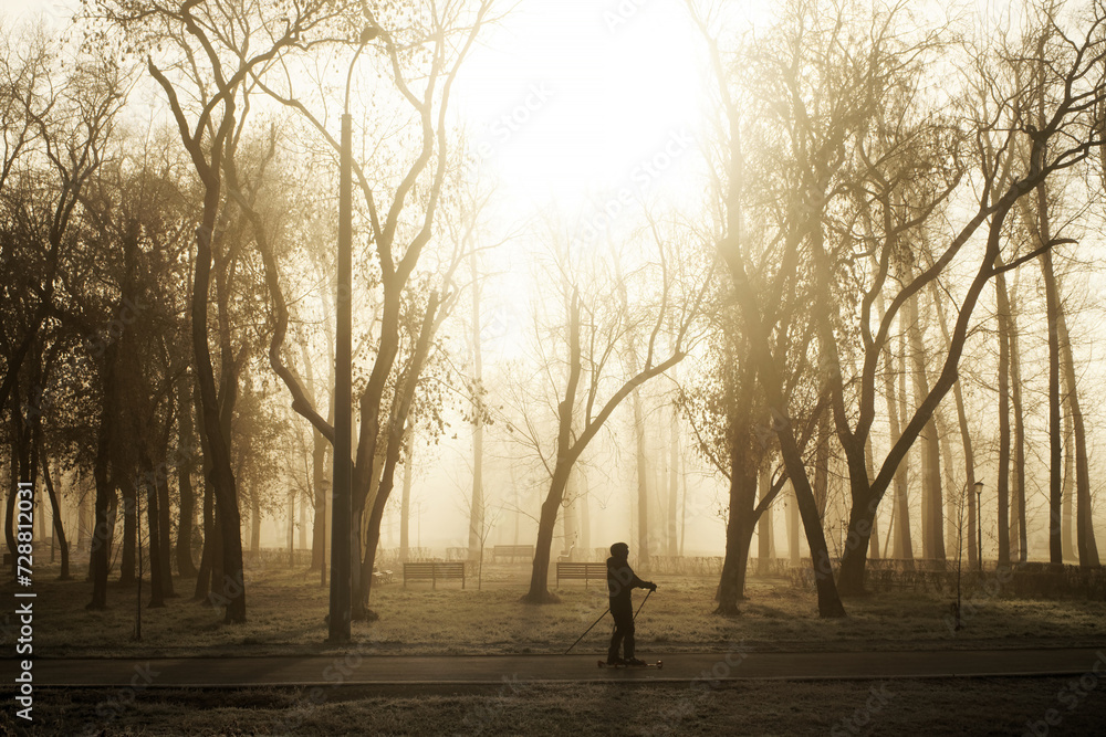 An athlete on roller skis warming up in a fall city park with morning mist, trees benches and lanterns. Photo. Contrasting natural light