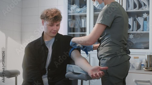 Professional nurse ties tourniquet carefully to prepare blond man arm for blood donation. Young woman cleans arm of patient with antiseptic wipe ensuring sterile site for blood collection photo