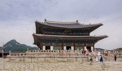 Gyeongbokgung Palace during cloudy summer day. With a girl in traditional clothing sitting on a stairs and tourists walking around.