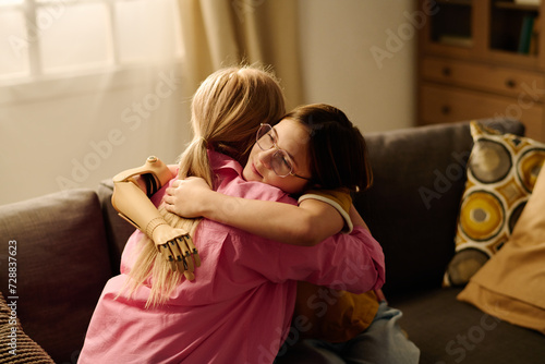 Young blond single mother and happy affectionate girl with myoelectric hand sitting in embrace on couch in living room photo