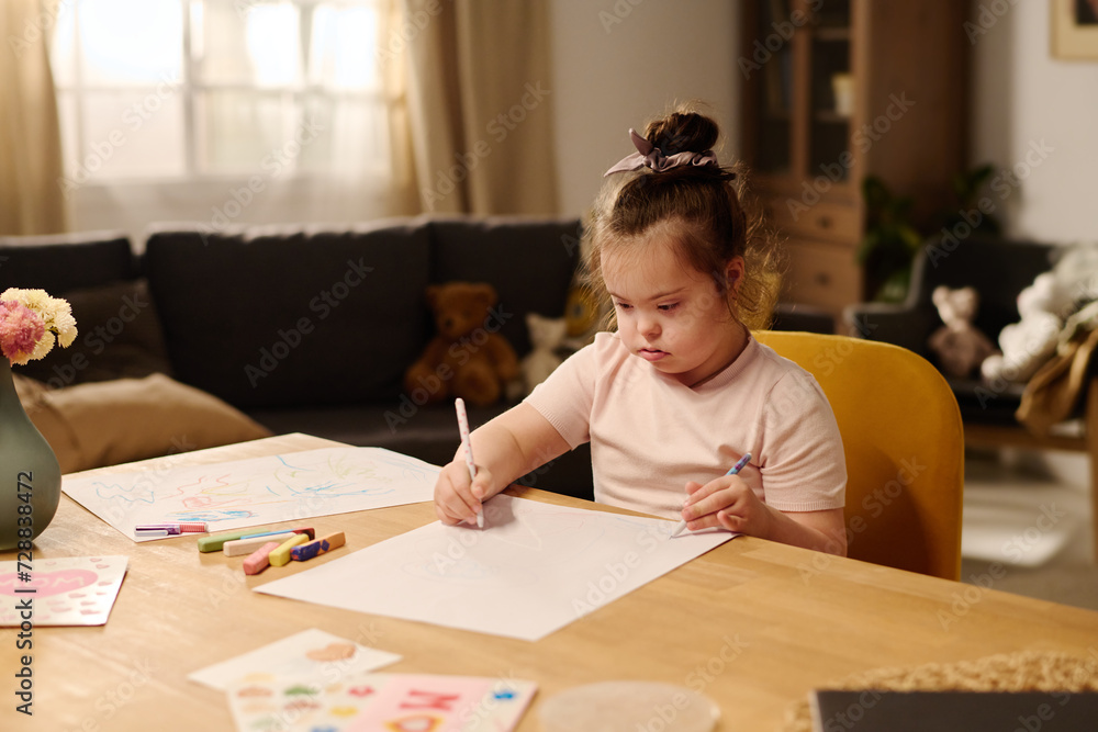 Cute little girl with mental disability sitting by table in front of camera and drawing with crayons and highlighters on sheet of paper Cute little girl with mental disability sitting by table in front of camera and drawing with crayons and highlighters on sheet of paper