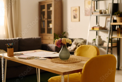 Wooden table with papers, crayons and grey vase with flowers standing in front of camera in the center of spacious living room