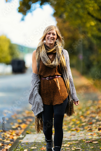 A young, blonde woman in a knitted look walks towards the camera along a sidewalk covered with autumn leaves.