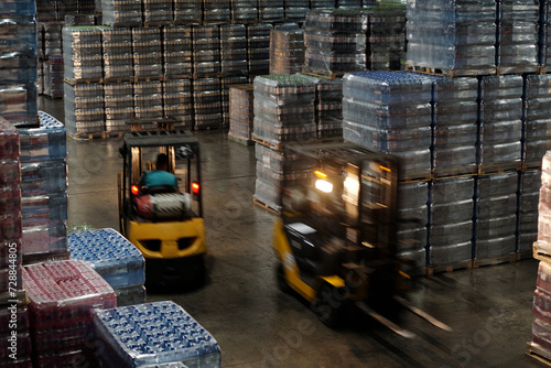 Blurry motion of two forklifts along aisle between huge stacks of packed plastic bottles containing mineral water, soda or some other drinks photo