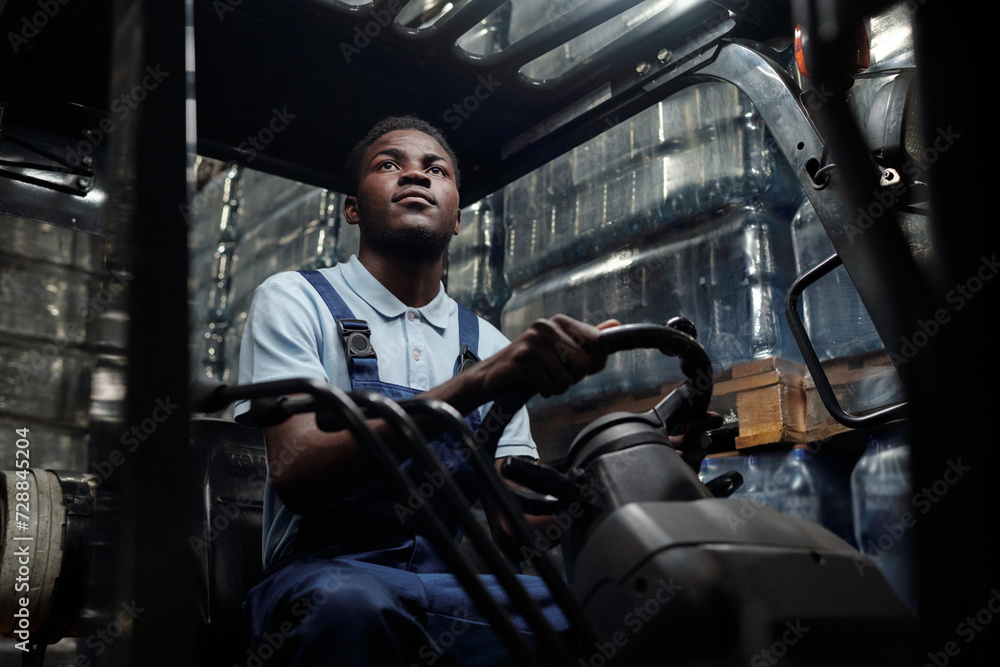 Young worker of huge industrial warehouse in coveralls sitting in forklift and driving it while moving along aisle between stacks of goods Young worker of huge industrial warehouse in coveralls sitting in forklift and driving it while moving along aisle between stacks of goods