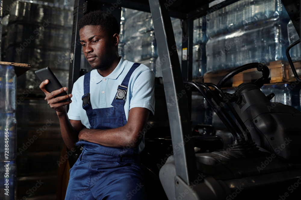 Young factory engineer sitting against door of forklift at break and texting in cellphone in distribution warehouse with stacks of bottled water Young factory engineer sitting against door of forklift at break and texting in cellphone in distribution warehouse with stacks of bottled water