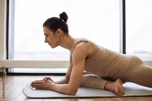 Calm caucasian brunette in beige sportswear lying on yoga mat stretching legs and hands in asana, panoramic windows in bright fitness center on background.